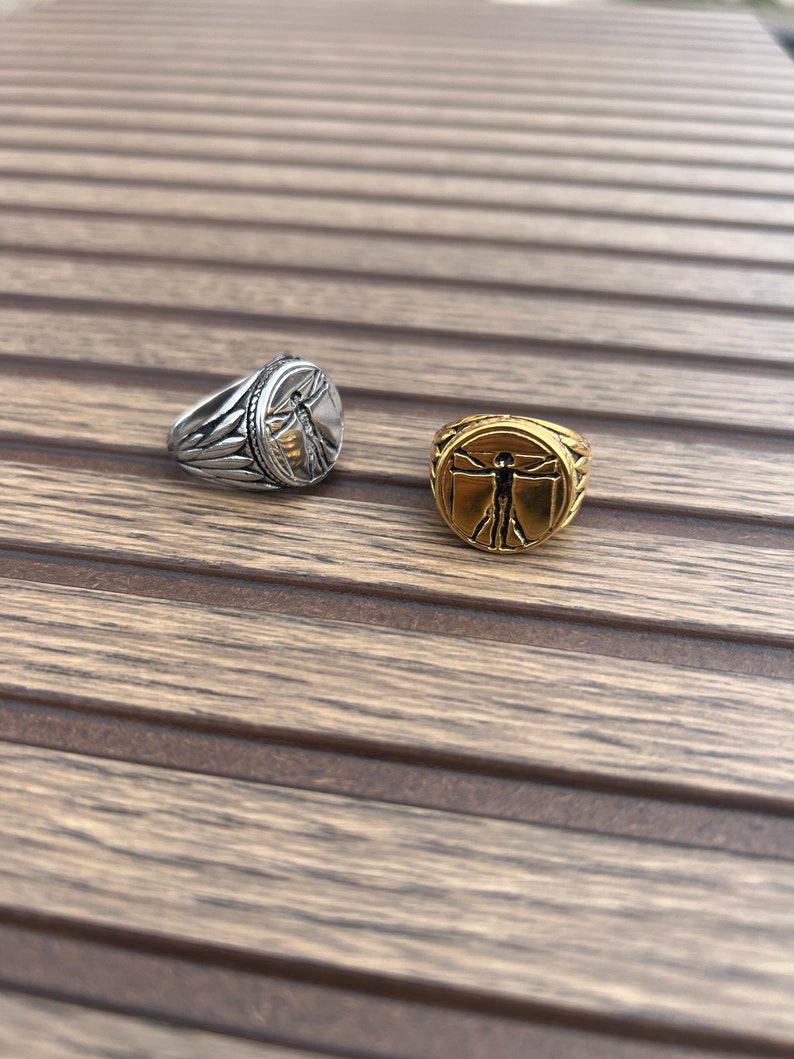 May include: Two silver and gold rings with a raised image of a man with outstretched arms. The rings are on a wooden surface.