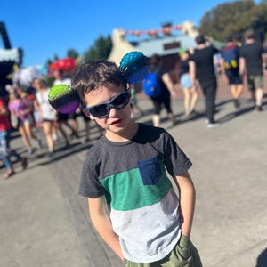 May include: A young boy wearing a green, white, and gray striped t-shirt with a blue pocket and green shorts. He is wearing sunglasses and has a pair of colorful mouse ears on his head.