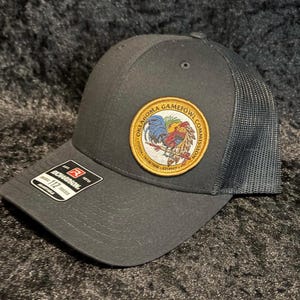 Puede incluir: Gorra de camionero gris oscuro con parte trasera de malla y visera curva. La gorra presenta un parche circular con las palabras "Oklahoma Gamefowl Commission" y un diseño de gallo colorido. Una pequeña etiqueta en la visera dice "Richardson 112 Trucker".