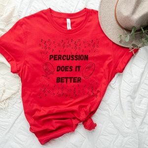 Può includere: Maglietta rossa con il testo "PERCUSSION DOES IT BETTER" in nero. La maglietta è decorata con note musicali e un tamburo.