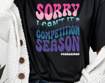 May include: Black t-shirt with the text "SORRY I CAN'T IT'S COMPETITION SEASON" in pink, blue, and purple. The hashtag #CHEERMOM is below the text. A white cardigan is worn over the shirt.