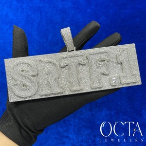 May include: A silver pendant featuring the word "SRITI" in large, raised letters, fully encrusted with sparkling diamonds. The pendant is held in a black gloved hand.