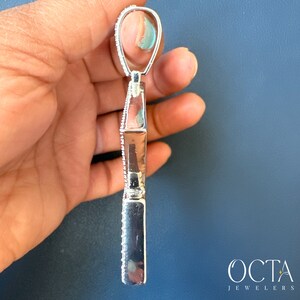 Puede incluir: Un colgante de plata con un dise&ntilde;o geom&eacute;trico, que presenta una fila de peque&ntilde;as piedras transparentes alrededor del per&iacute;metro. El colgante se sostiene en una mano, con la parte posterior del colgante mirando hacia la c&aacute;mara.