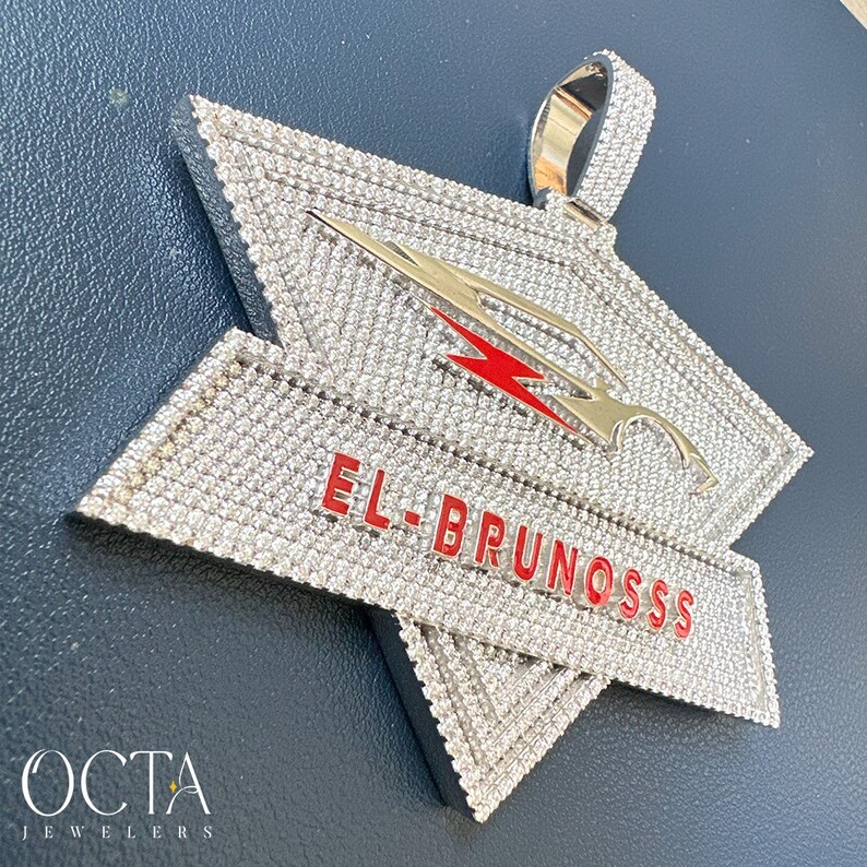 Puede incluir: Un colgante de plata con un dise&ntilde;o en forma de estrella, cubierto de piedras de circonita c&uacute;bica transparentes. El colgante presenta un rayo rojo y el texto "EL-BRUNOSSS" en rojo.