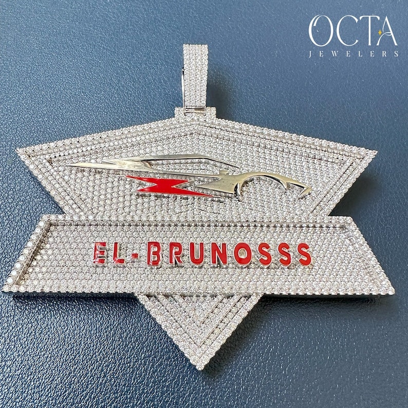 Puede incluir: Un colgante de plata con un dise&ntilde;o engastado en diamantes que presenta un coche estilizado y el texto "EL BRUNOSSS" en letras rojas. El colgante es una pieza llamativa con una est&eacute;tica moderna y lujosa.