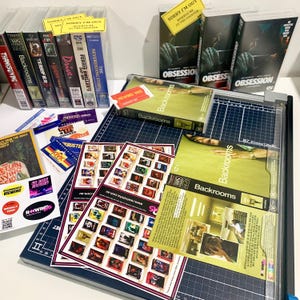 May include: A collection of VHS tapes including titles like Backrooms, Terrifier, and Obsession, alongside printed movie posters, membership cards, and stickers, all arranged on a dark blue cutting mat with grid lines and a clear plastic trimmer.
