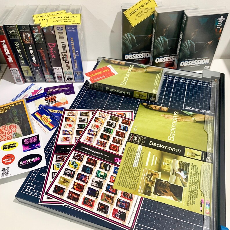 May include: A collection of VHS tapes including titles like Backrooms, Terrifier, and Obsession, alongside printed movie posters, membership cards, and stickers, all arranged on a dark blue cutting mat with grid lines and a clear plastic trimmer.