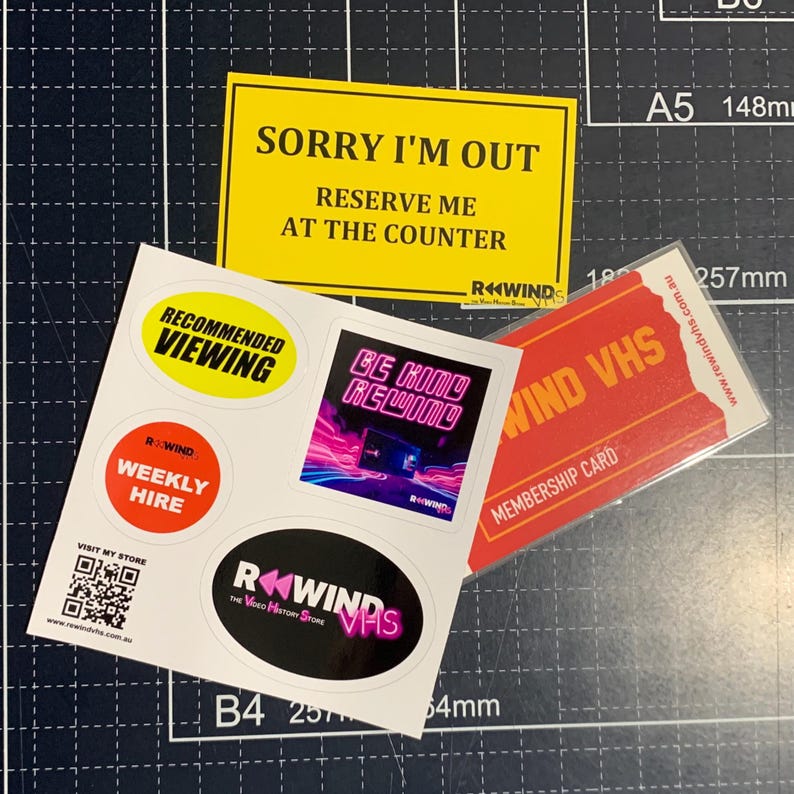 May include: A collection of stickers and a membership card featuring the Rewind VHS branding. The items include a yellow rectangular sign, a white sticker sheet with various circular and square decals, and a red rectangular membership card.