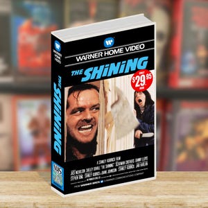 Puede incluir: Una cinta VHS de la película "The Shining" con Jack Nicholson en la portada. La carátula muestra a Nicholson y a una mujer con un cuchillo. La cinta está etiquetada como "Warner Home Video".