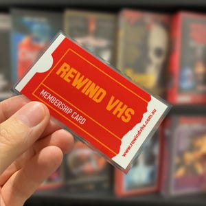 May include: A rectangular membership card with a red background and white borders. The text REWIND VHS is printed in large orange letters, with MEMBERSHIP CARD in white below it. The website www.rewindvhs.com.au is printed in red along the side.