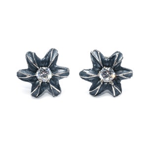 Puede incluir: Un par de pendientes de plata en forma de flor con un brillante transparente en el centro de cada flor. Los pendientes tienen un acabado oxidado gris oscuro.