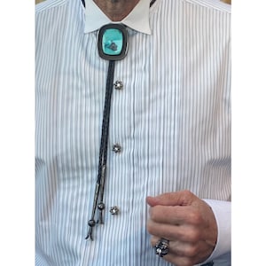 Może przedstawiać: Turkusowy krawat bolo z srebrnym oprawą i czarnym plecionym sznurkiem. Krawat bolo jest noszony na białej koszuli w paski.