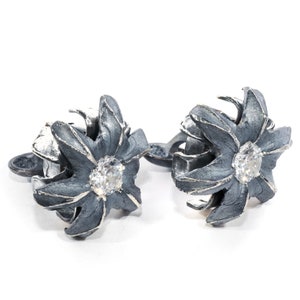 Może przedstawiać: Para srebrnych kolczyków z kwiatowym wzorem. Każdy kolczyk ma srebrny kwiat z przezroczystym kryształem w środku. Kwiaty są wykonane z fakturowanego metalu.