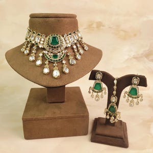 Puede incluir: Un collar tipo gargantilla en tono dorado con piedras verde esmeralda y cristales transparentes, acompañado de pendientes a juego y un adorno para la cabeza maang tikka. El conjunto presenta metalistería intrincada y detalles colgantes.