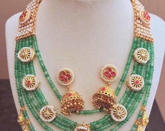 May include: A multi-strand necklace set with green beads, gold accents, and red and white stones. The necklace has a layered design with dangling charms. The set also includes matching earrings.
