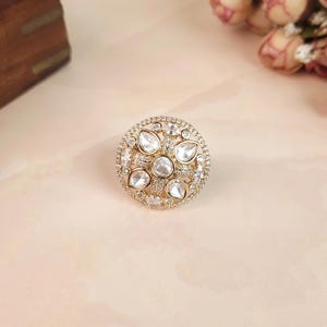 May include: A circular gold-tone ring featuring a central round clear stone surrounded by four pear-shaped clear stones, all set within a frame encrusted with numerous tiny clear pavé stones arranged in a detailed, symmetrical floral-inspired pattern.