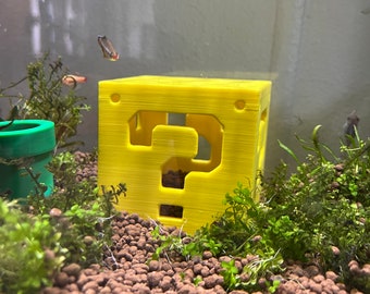 May include: A yellow, 3D-printed question mark block, a recognizable element from a popular video game, is placed in an aquarium. The aquarium also contains aquatic plants, gravel, and small fish. A green pipe is visible in the background.