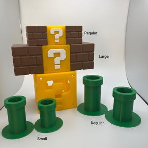 May include: A collection of 3D-printed items inspired by a video game. Includes a yellow question mark block, brown brick blocks, and green pipes in various sizes. The text "Regular", "Large", and "Small" are visible.