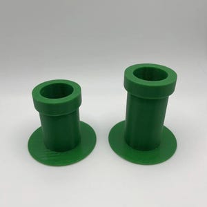 May include: Two green plastic pipes, resembling those from a video game, are displayed against a white background. Each pipe has a circular base and a cylindrical body with a circular opening at the top. The pipes are a vibrant shade of green.
