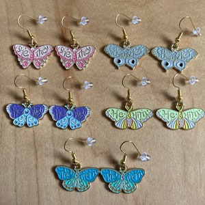 May include: A set of ten butterfly earrings with different colors and designs. The earrings feature the words "She They", "She Her", "He Him", and "They Them" in a whimsical font.