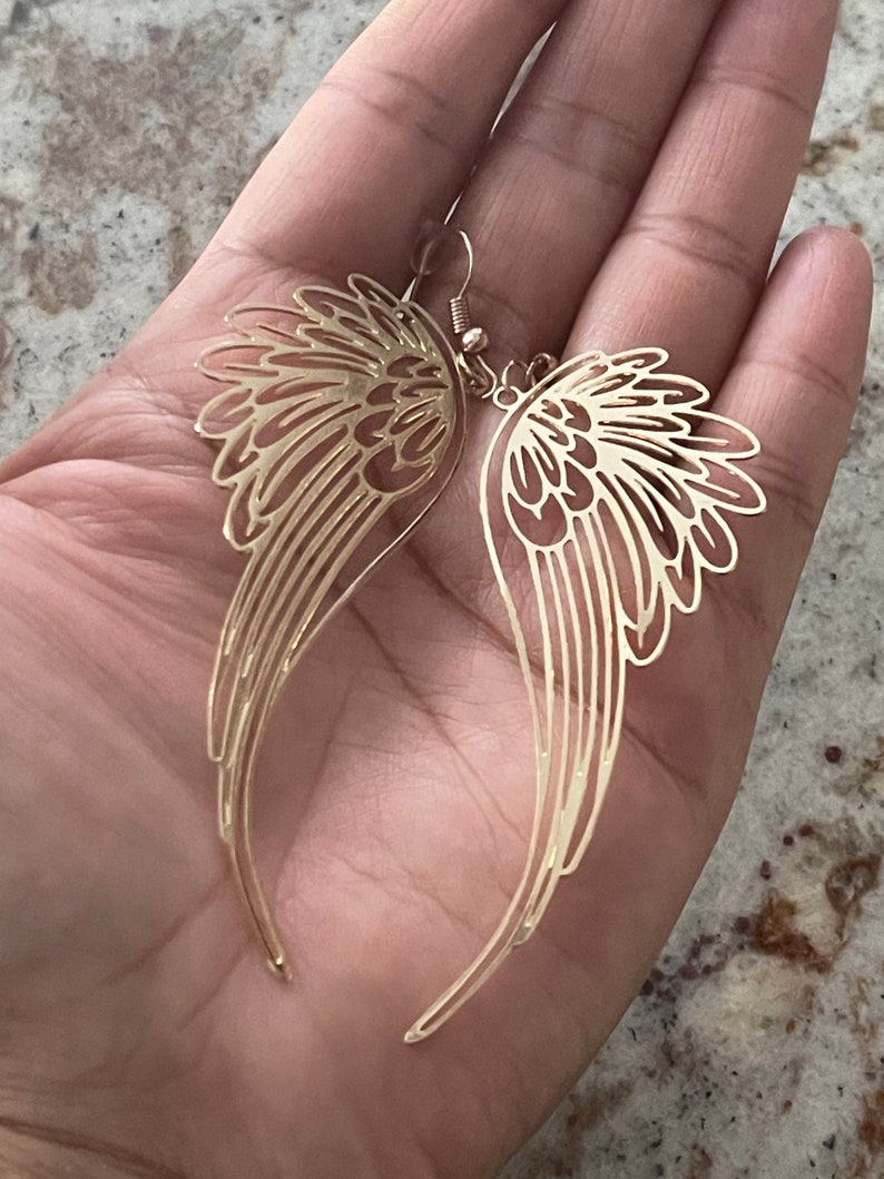 Puede incluir: Un par de pendientes colgantes de tono dorado con un delicado dise&ntilde;o calado de alas de &aacute;ngel.