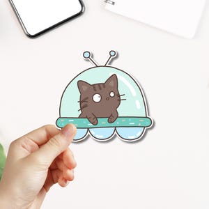 May include: A brown cat with big eyes sits in a light blue and green spaceship with a white outline. The spaceship has a small antenna on top.