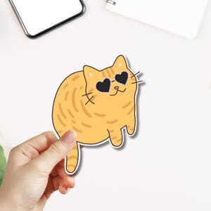 Könnte beinhalten: Eine orangefarbene Comic-Katze mit Herz-Sonnenbrille. Die Katze sitzt mit den Beinen unter dem Körper und hat einen glücklichen Gesichtsausdruck.