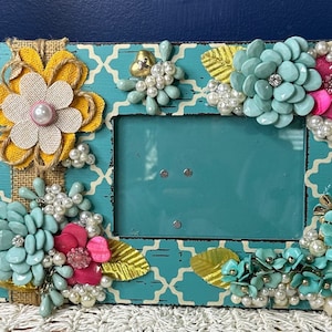 May include: A turquoise picture frame decorated with faux flowers and pearls. The frame has a white and turquoise geometric pattern.