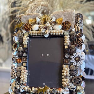 May include: A picture frame decorated with a variety of materials, including pearls, shells, animal print fabric, and rhinestones. The frame is a dark brown color and has a rectangular shape.