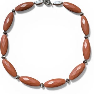 May include: A necklace featuring faceted, oval-shaped beads in a muted coral color. The beads are strung together with small, silver-toned metal accents and a decorative clasp. The necklace is designed to be worn around the neck.