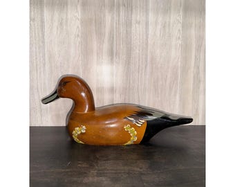 May include: A wooden duck figurine with a brown body, black tail, and a green and black beak. The duck has painted details, including white and yellow floral accents on its sides. The figurine is a decorative item.