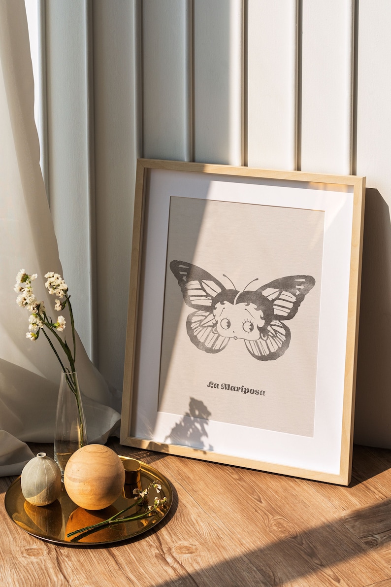 May include: A framed print of a black and white butterfly with a cartoon face. The print is on a beige background and the text "La Mariposa" is printed below the butterfly.