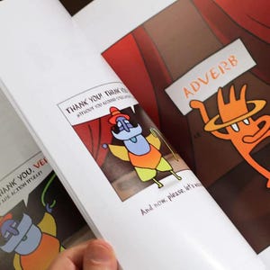 A hand flipping through The Language Theater book, showing a colorful adverb character on a stage-themed page with bold illustrations.