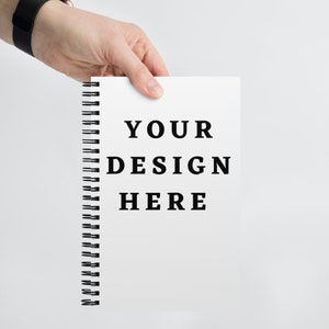 Könnte beinhalten: Ein weißes Spiralnotizbuch, das von einer Hand gehalten wird, mit dem Text "YOUR DESIGN HERE" in Schwarz gedruckt. Die Seiten des Notizbuchs sind leer und bieten Platz für individuelle Designs oder Notizen. Die Spiralbindung ist schwarz.