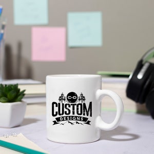 May include: White ceramic mug with a black and white logo that says "C-D Custom Designs" with a mountain range and trees in the background.