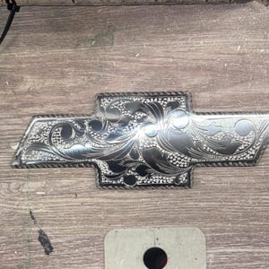 May include: Silver-colored Chevrolet emblem with intricate floral engraving. The emblem features a classic bow tie design, with detailed patterns across its surface. The metal has a polished finish, reflecting light. The emblem is likely for a vehicle.
