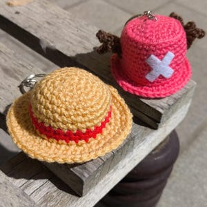 May include: Two crochet keychains shaped like hats. One is tan with a red band, the other is pink with a white cross and brown antlers. Both are sitting on a weathered wooden surface.