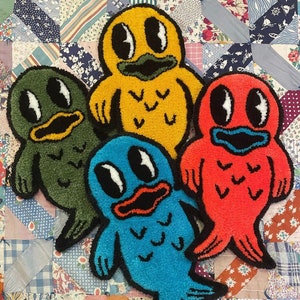 May include: Four colorful, whimsical rug-like fish with embroidered details. The fish are yellow, green, blue, and red. Each fish has a unique design and features a smiling face with large eyes.