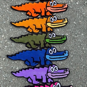 May include: A collection of seven tufted textile rugs shaped like cartoon crocodiles, arranged in a vertical column. Each rug features a distinct solid color including dark red, orange, yellow-orange, olive green, blue, purple, and bright pink.