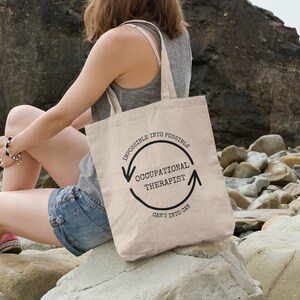 Peut inclure: Un sac fourre-tout en toile beige avec du texte noir qui dit "Impossible into Possible Occupational Therapist Can't into Can". Le texte est dans un cercle avec des flèches pointant dans des directions opposées.