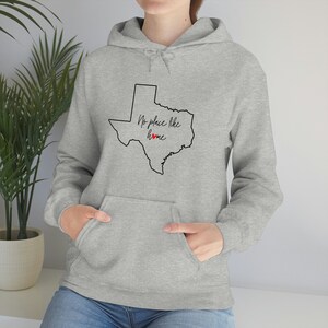May include: A light gray hoodie with a front pocket and a drawstring hood. The hoodie features a black outline of the state of Texas with the words "No place like home" and a red heart.