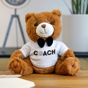 Puede incluir: Un oso de peluche marrón que lleva una camisa blanca con una pelota de béisbol y la palabra "COACH" impresa en ella. El oso lleva una pajarita negra.