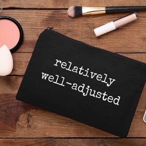 May include: Black makeup bag with a white zipper and the text "relatively well-adjusted" printed on the front. The bag is lying on a wooden surface with other makeup items.