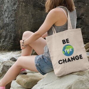 Può includere: Una persona seduta su delle rocce vicino al mare che tiene una borsa di tela beige con il testo "BE THE CHANGE" e un globo terrestre verde e blu.