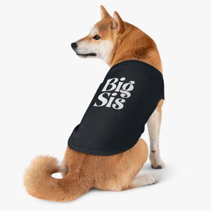 Op de afbeelding: Een bruine en witte hond die een zwart tanktop draagt met de tekst "Big Sis" in wit. De hond zit en kijkt naar links.