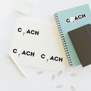 May include: Four white golf ball stickers with the word "COACH" in black text. The stickers are on a light beige background with a white pen and paper clips.
