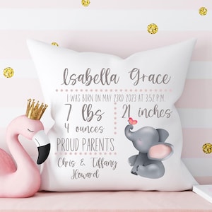 以下が含まれることがあります： 「Isabella Grace」の名前と誕生日の詳細が記載された白い装飾枕。2023年5月23日、3.18 kg、53.34 cm。ピンクの耳とハートを持つ灰色の象が特徴です。「誇り高き両親：Chris & Tiffany Howard」というテキストが含まれています。