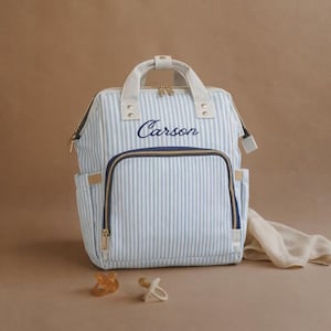 May include: A blue and white striped baby changing bag with the name "Carson" embroidered in navy blue. The bag has a front pocket, side pockets, and a top handle. Two dummies and a muslin cloth are next to the bag.