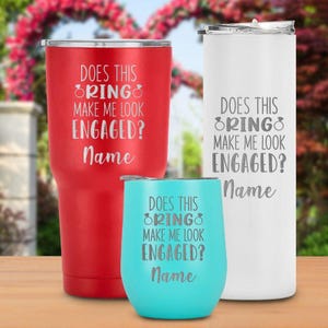 May include: Three stainless steel tumblers in red, white, and teal. Each tumbler has the text "DOES THIS RING MAKE ME LOOK ENGAGED? Name" engraved on the front. The tumblers are different sizes and shapes.