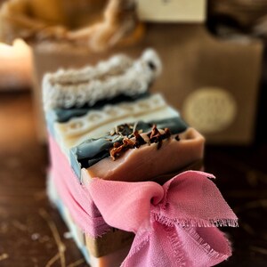 May include: A stack of handmade soaps in various colours, including blue, beige, and pink, tied with a pink ribbon. The soaps have textured surfaces and are decorated with dried botanicals. The background includes a blurred package and a tag.